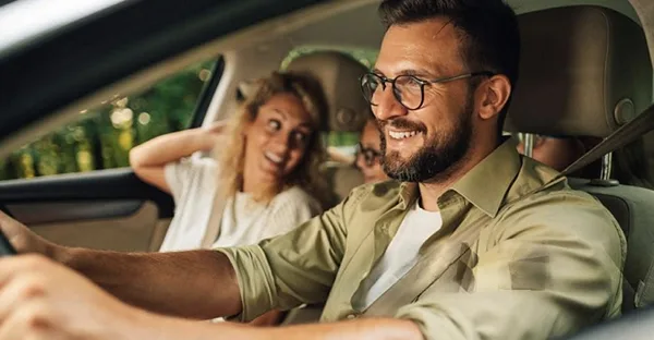 Ein lächelnder Mann mit Brille fährt Auto, während im Hintergrund eine fröhliche Frau als Beifahrerin zu sehen ist.