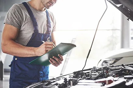 Ein Kfz-Mechaniker in blauer Arbeitslatzhose macht Notizen auf einem Klemmbrett, während er vor einem geöffneten Motorraum eines Fahrzeugs in einer hellen Werkstatt steht.