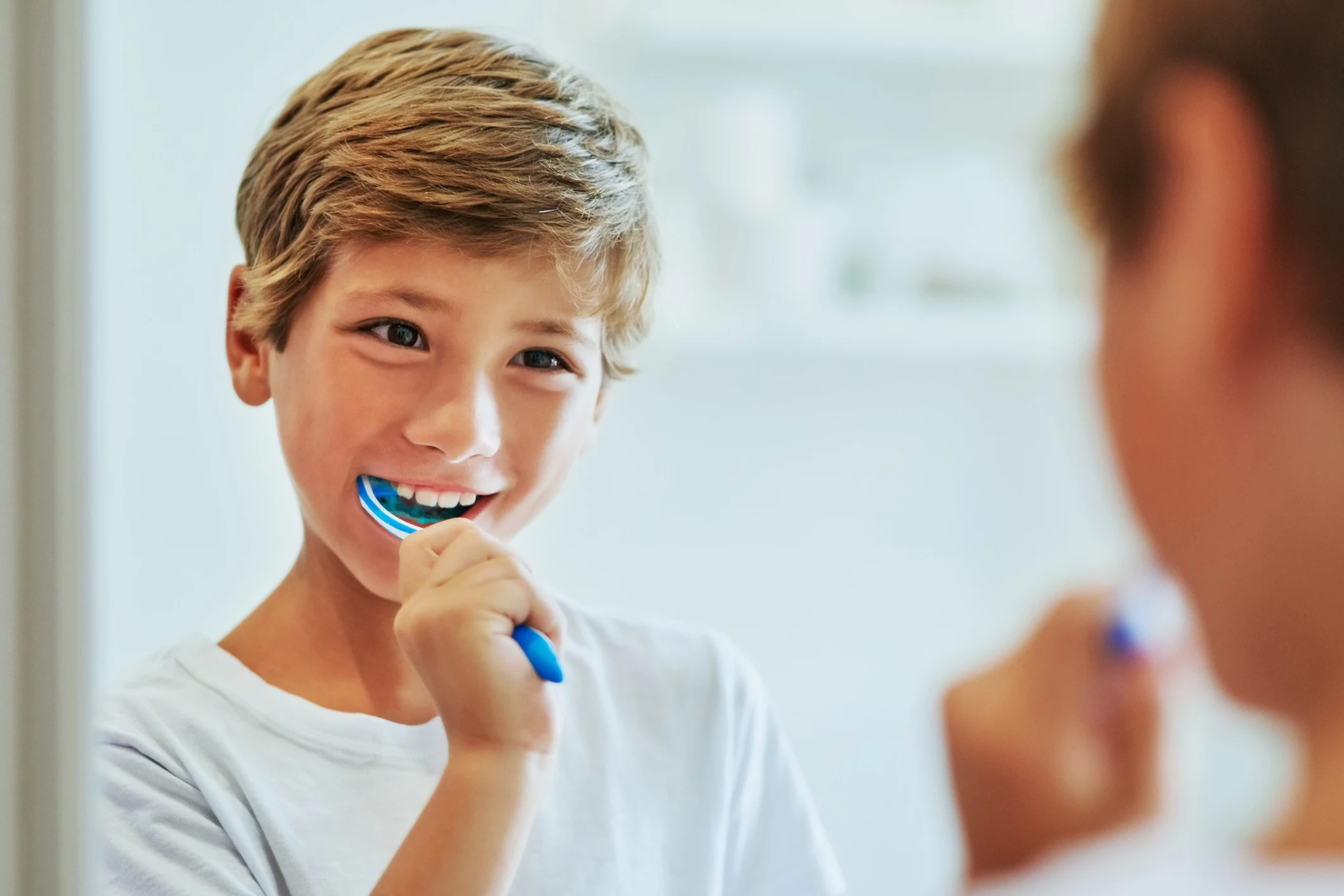 Ein lächelnder Junge mit blonden Haaren putzt sich vor dem Spiegel die Zähne mit einer blauen Zahnbürste im hellen Badezimmer.