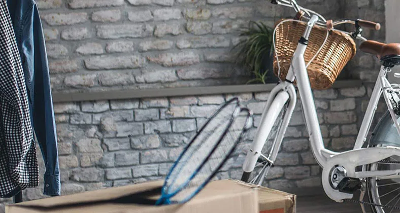 Weißes Fahrrad mit Weidenkorb am Lenker steht vor grauer Backsteinwand, daneben hängen Hemden und liegt ein Surfbrett auf einem Karton.