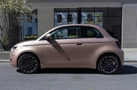 Ein rosafarbener Fiat 500 mit zweifarbigem Dach und schwarzen Felgen steht in der Seitenansicht vor einem modernen Gebäude mit Glasfassade.