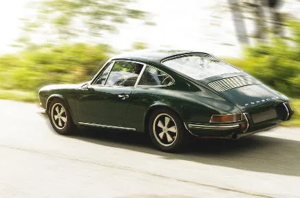 Ein dunkelgrüner klassischer Porsche 911 fährt dynamisch auf einer Landstraße, umgeben von verschwommener grüner Vegetation im Hintergrund.