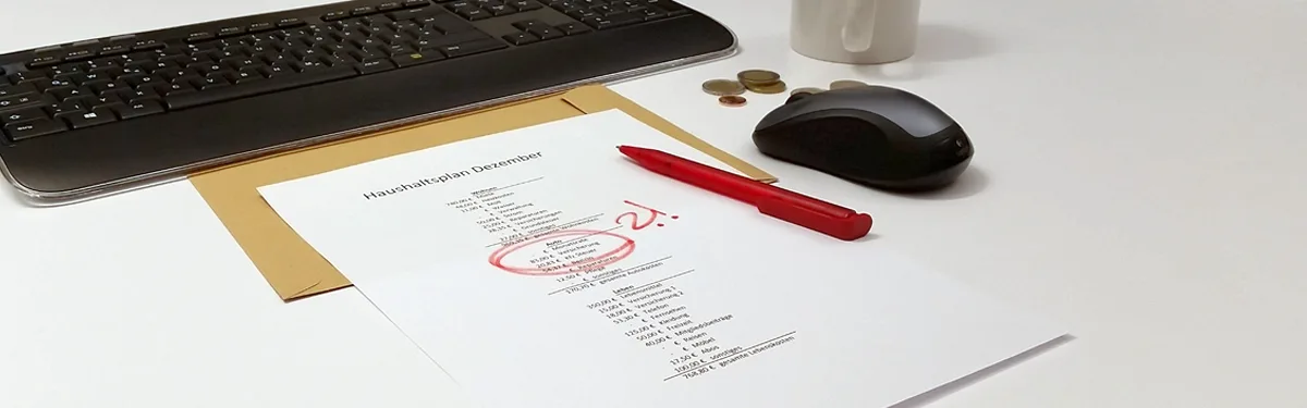 weiße Tischplatte, auf der folgende Gegenstände abgestellt sind: Tastatur, Maus, weiße Tasse, Münzgeld, ein DIN/A4 Blatt, das einen braunen Umschlag überdeckt, auf dem Blatt ist ein Haushaltsplan für Dezember abgebildet, roter Kugelschreiber