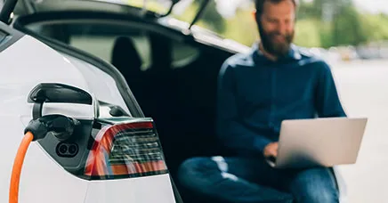 Mann mit Laptop sitzt im Kofferraum eines Elektroautos, das über ein orangefarbenes Kabel aufgeladen wird.