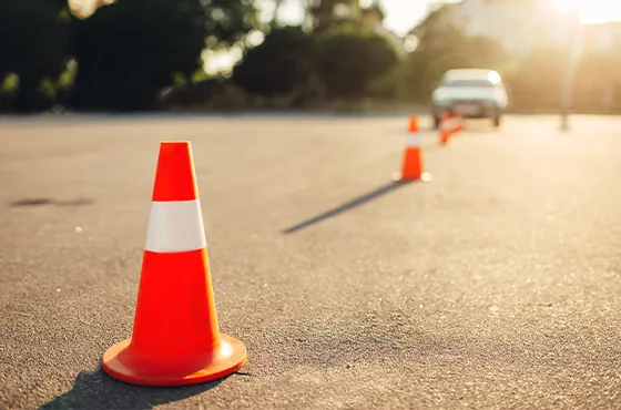 Orange Verkehrskegel auf einer Straße im Sonnenlicht, mit weiteren Kegeln und einem Auto im unscharfen Hintergrund.