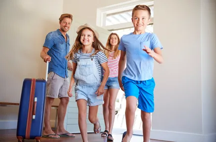 Eine vierköpfige Familie betritt fröhlich ihr neues Zuhause mit Gepäck, die Kinder laufen voraus durch die helle Eingangstür.