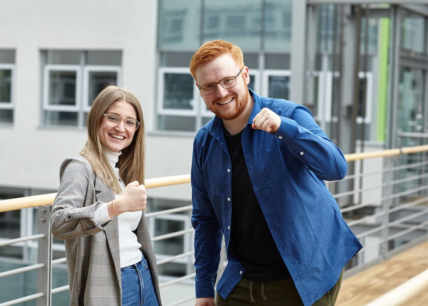 Zwei lächelnde junge Berufstätige zeigen Daumen hoch vor einem modernen Bürogebäude mit Glasfassade.