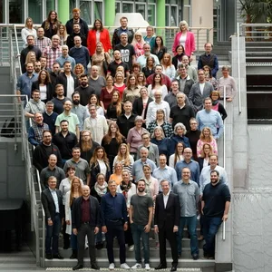 Großes Gruppenfoto von etwa 80-100 Personen auf einer Außentreppe vor einem modernen Gebäude, aufgenommen von erhöhter Position.