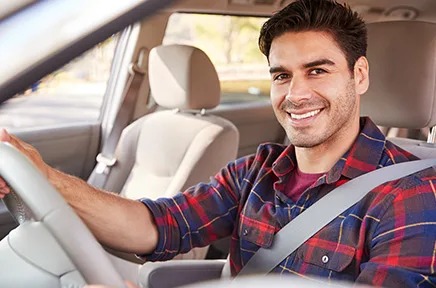Ein lächelnder junger Mann mit kariertem Hemd sitzt angeschnallt am Steuer eines Autos und blickt freundlich zur Seite.