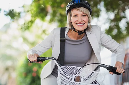 Lächelnde Frau mit Fahrradhelm und grauem Blazer steht mit ihrem Fahrrad mit Korb in einem sonnigen Park.