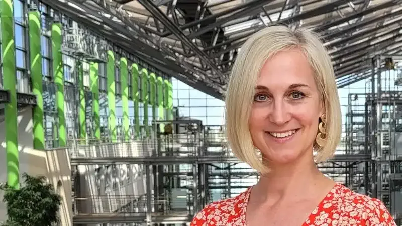 Blonde Frau in rotem Blumenkleid lächelt in moderner Architektur mit Glasfassade, grünen Akzenten und industriellem Stahlgerüst im Hintergrund.