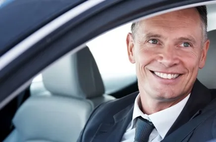 Lächelnder Geschäftsmann in Anzug und Krawatte sitzt am Steuer eines Autos und blickt freundlich durch das geöffnete Seitenfenster.