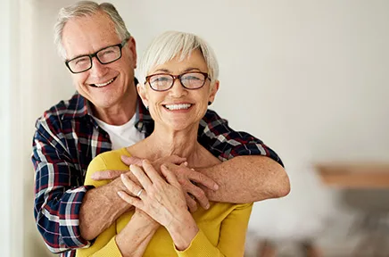 Ein älteres Paar mit Brillen lächelt glücklich in die Kamera, während der Mann die Frau von hinten umarmt, beide tragen lässige Kleidung in einem hellen Raum.