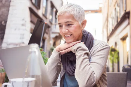 Lächelnde Frau mit kurzen grauen Haaren arbeitet an einem Laptop in einem Straßencafé im Freien.