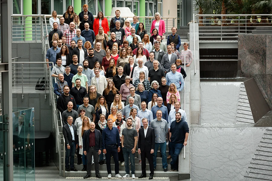 Großes Gruppenfoto von etwa 80-100 Personen auf einer Außentreppe vor einem modernen Gebäude, aufgenommen von erhöhter Position.