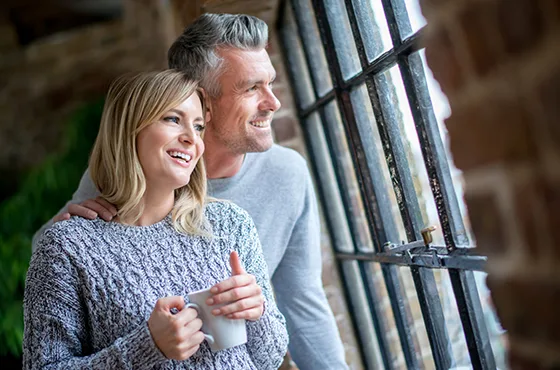 Lächelndes Paar in Strickpullovern schaut gemeinsam durch ein großes Fenster, die Frau hält eine Tasse.