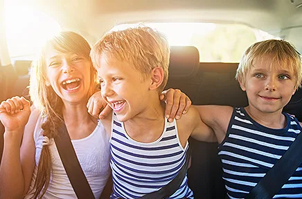 Drei fröhliche Kinder sitzen angeschnallt auf der Rücksitzbank eines Autos und lachen gemeinsam, während Sonnenlicht durch die Scheibe fällt.