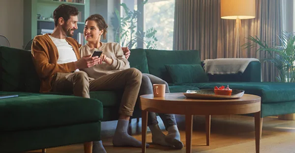 Ein Paar sitzt entspannt auf einem grünen Sofa in einem modern eingerichteten Wohnzimmer und schaut gemeinsam auf ein Smartphone.