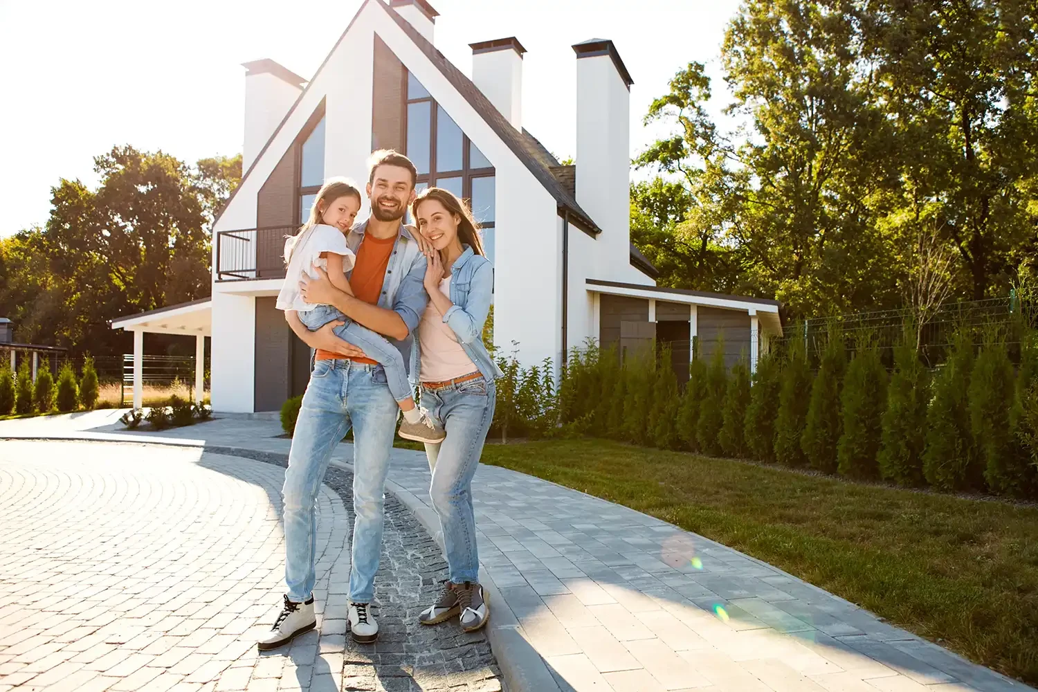 Eine junge Familie mit Kind steht glücklich vor ihrem modernen Einfamilienhaus mit weißer Fassade und großen Fenstern im sonnigen Garten.