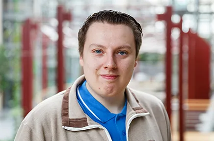 Porträtfoto einer Person mit kurzen braunen Haaren in beiger Jacke und blauem Shirt, die freundlich in die Kamera lächelt, vor einem hellen Bürogebäude mit roten Säulen im Hintergrund.