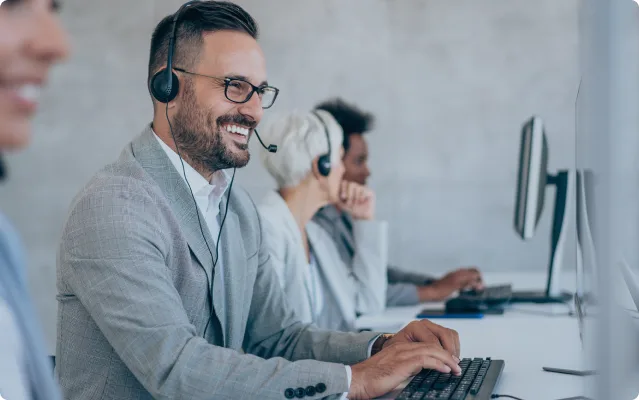 Lächelnder Mann mit Headset und Brille arbeitet an einem Computer in einem Callcenter, umgeben von Kollegen an ihren Arbeitsplätzen.
