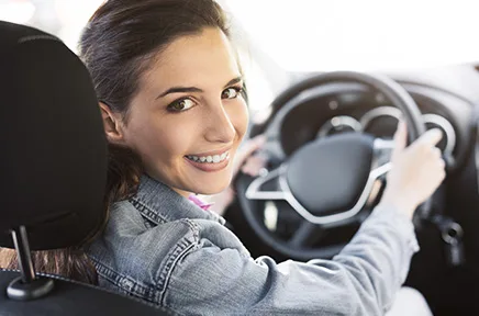 Lächelnde junge Frau mit Jeansjacke sitzt am Steuer eines Autos und blickt über ihre Schulter zur Kamera.