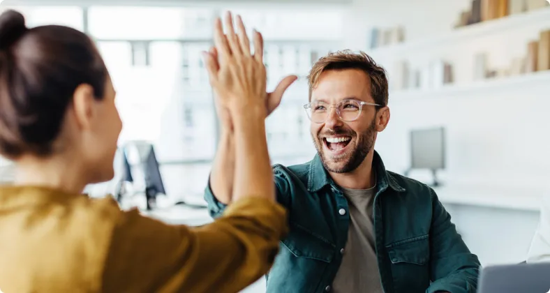 Zwei lächelnde Kollegen geben sich im hellen Büro ein High-Five, wobei der Mann eine Brille trägt und beide eine erfolgreiche Zusammenarbeit feiern.